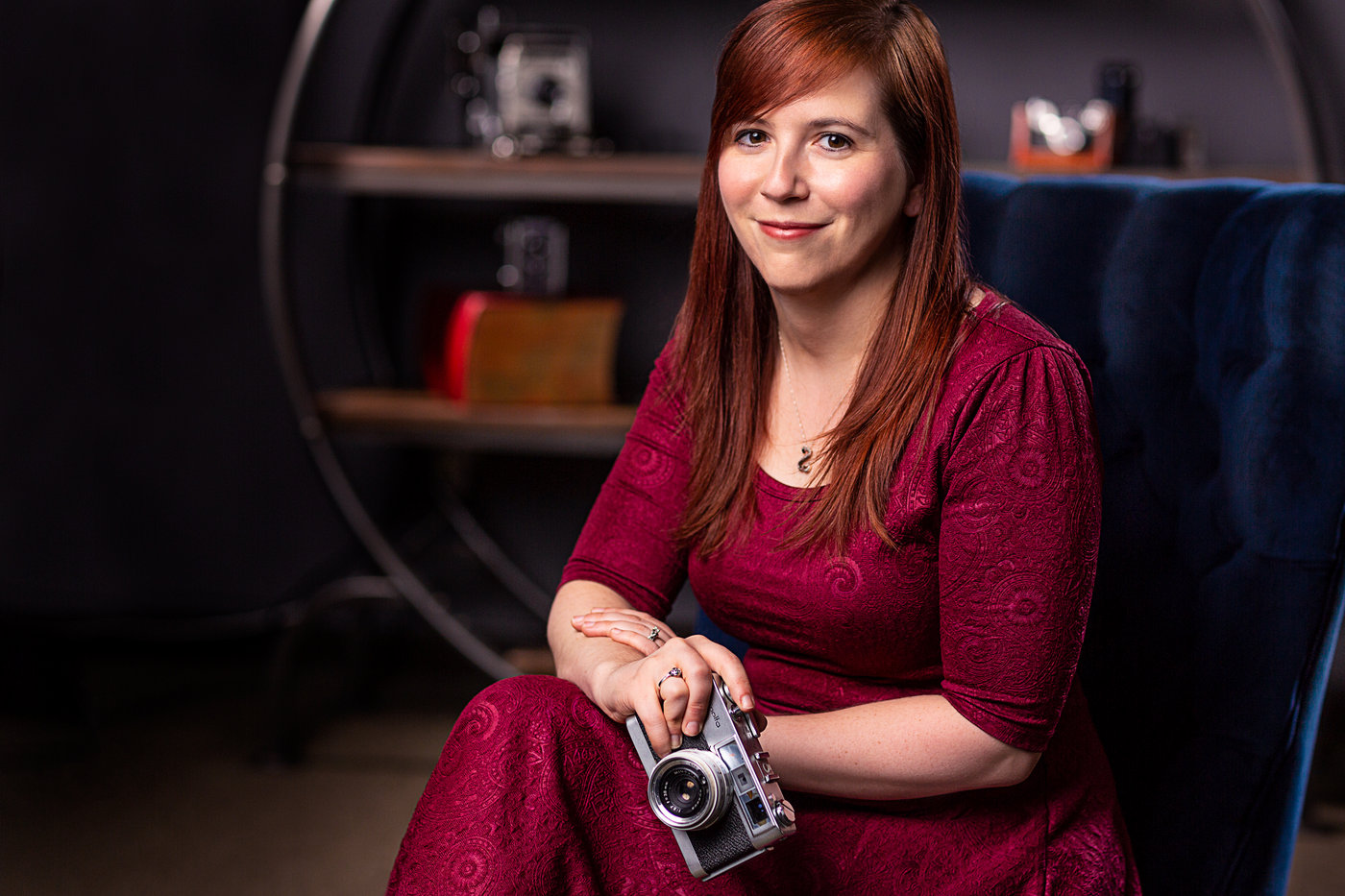 a woman poses with a vintage camera while wearing a dark red dress in front of a shelf of vintage cameras to demonstrate one of the tips for great branding photos