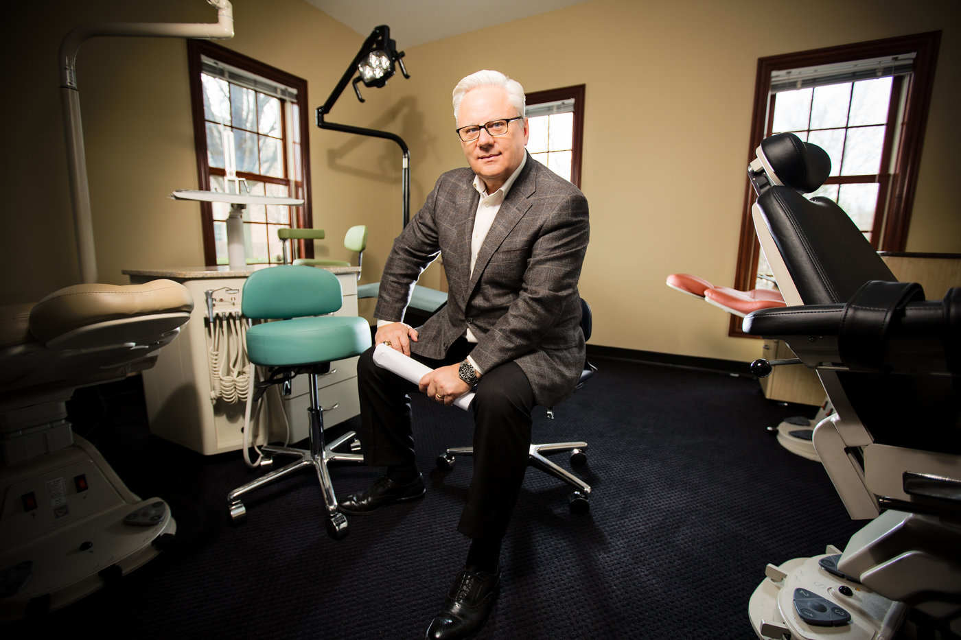 a man in a suit sits on a stool in a room full of dental equipment to demonstrate one of the tips for great brand photos