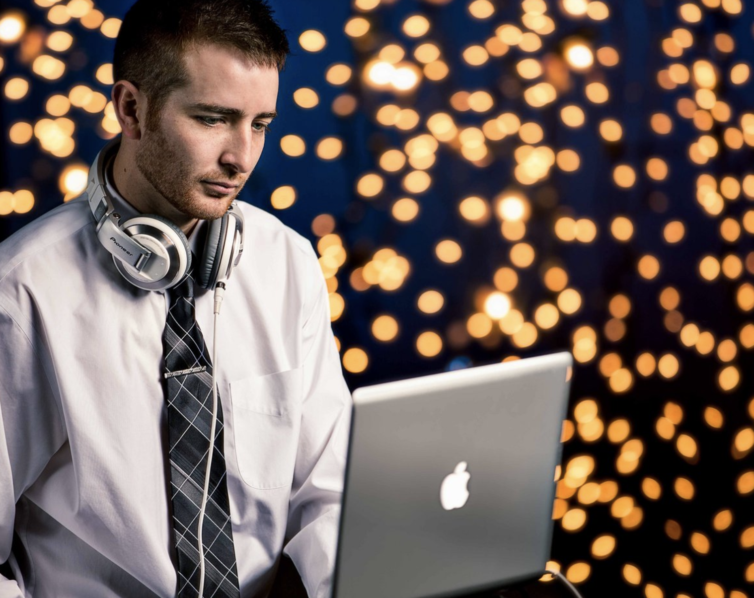 a DJ poses in front of a background full of string lights with a laptop and headphones to demonstrate one of the tips for great branding photos