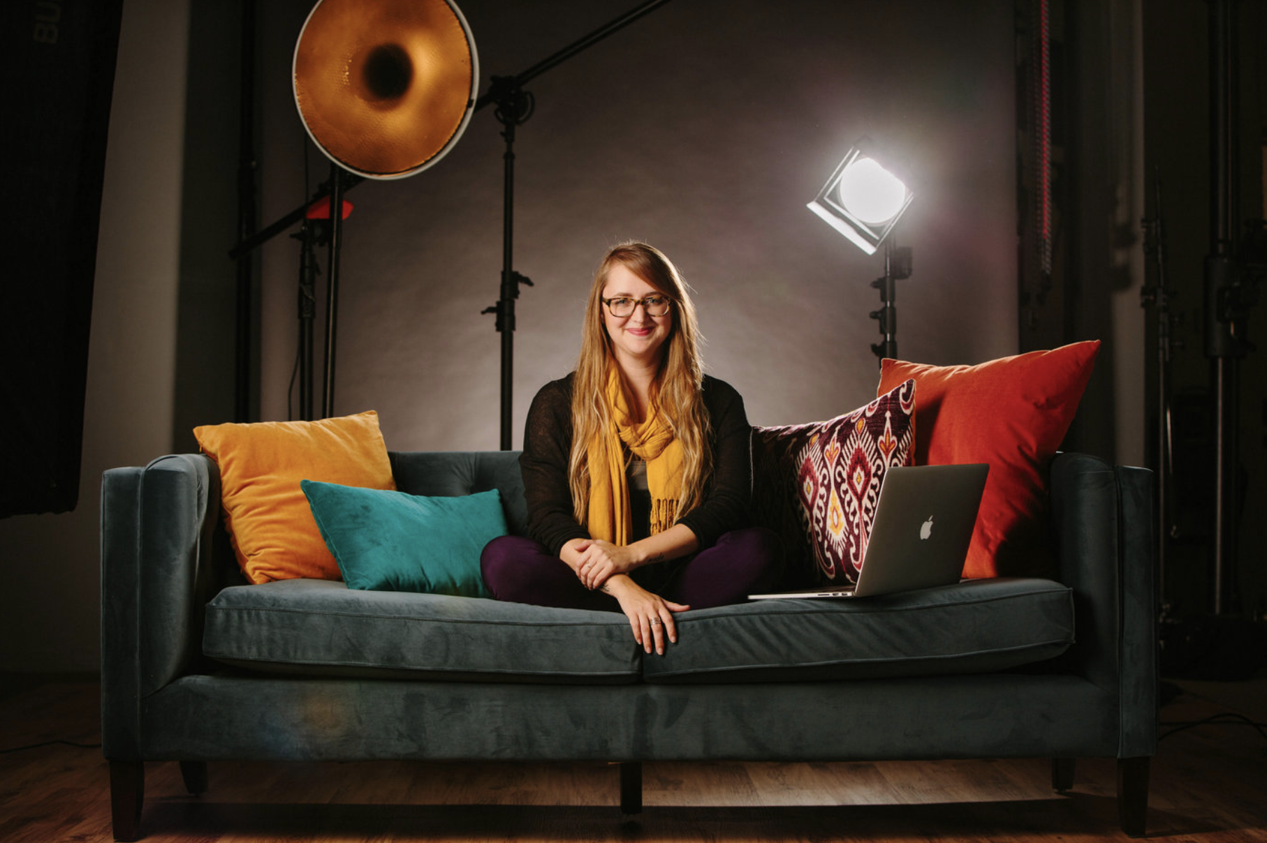 a blonde woman sits on a blue couch in a photo studio surrounded by colorful pillows to demonstrate one of the tips for great branding photos